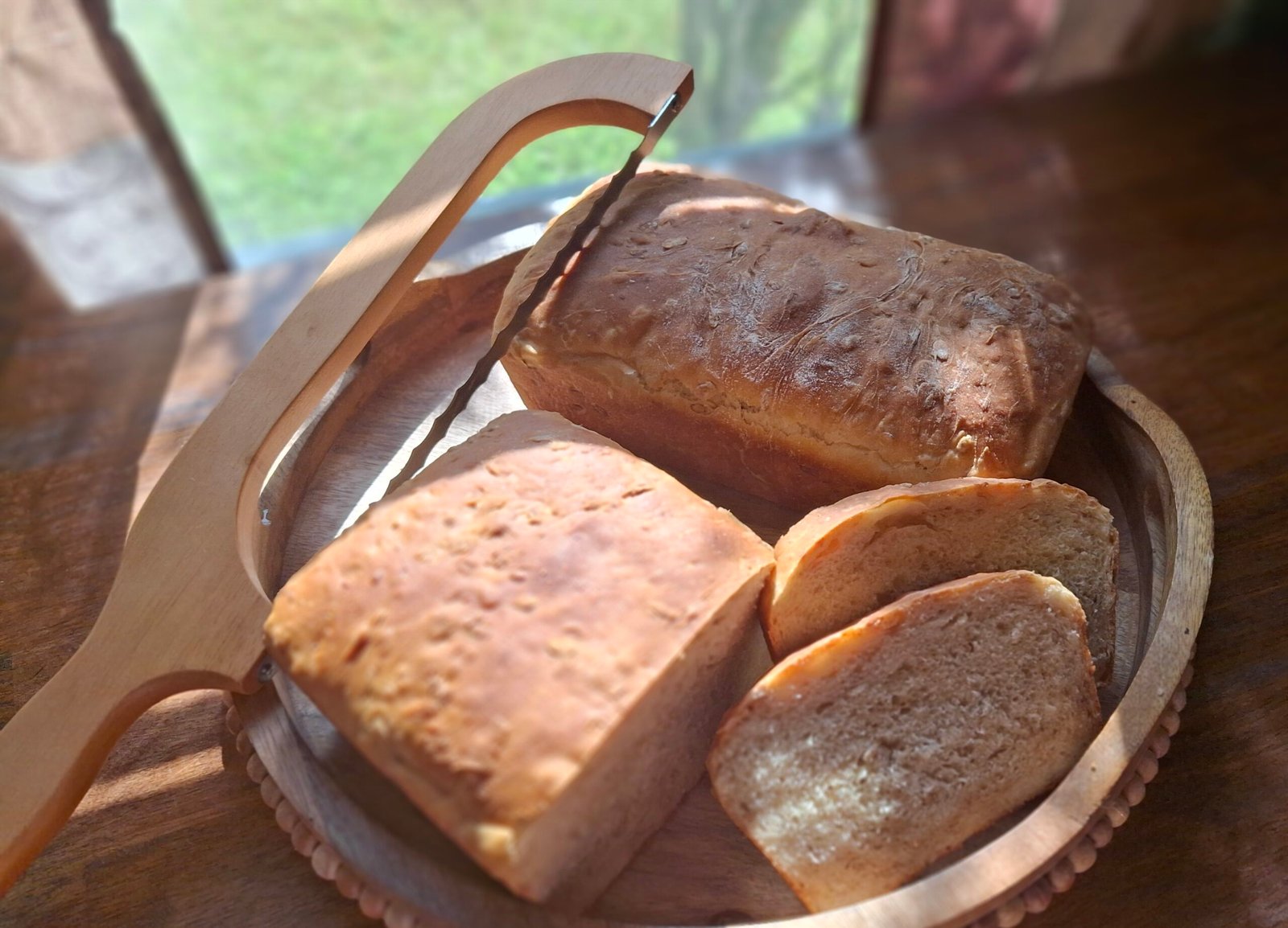 Two freshly baked homemade sandwich bread loaves on a round wooden serving board with a wooden bread slicer and several sliced pieces in warm natural sunlight.